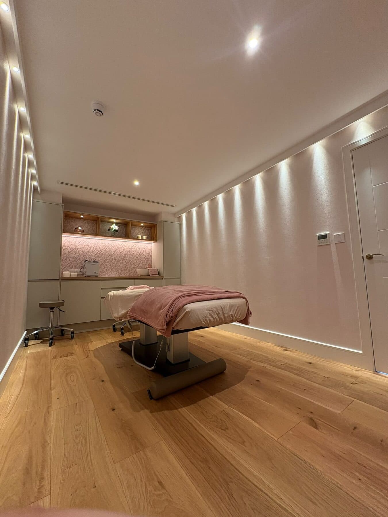 Treatment room with a run of downlights washing the wall, recessed cove lighting and lit display shelves over the basin